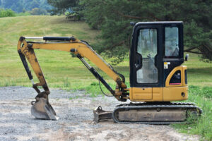 Compact yellow caterpillar excavator stands on gravel near grassy field. Construction equipment used for digging, earthworks, and small projects.